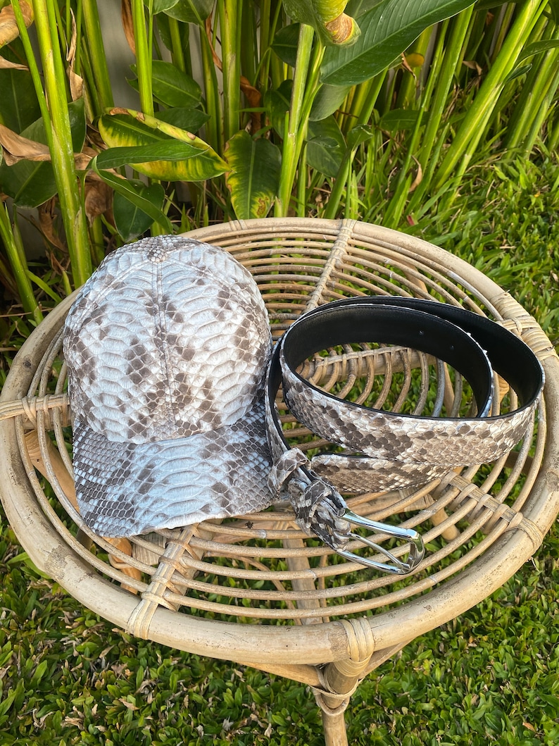 May include: A gray and white snakeskin baseball cap and a matching snakeskin belt with a silver buckle. The cap and belt are on a woven wicker table.