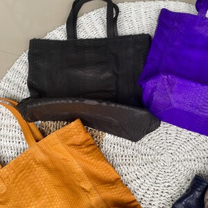 Puede incluir: Cuatro bolsos de mano diferentes hechos de piel de serpiente. Los bolsos son de color negro, morado, naranja y azul marino. Los bolsos est&aacute;n sobre una estera blanca tejida.