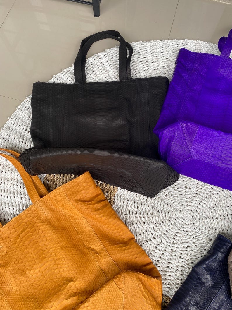 Puede incluir: Cuatro bolsos de mano diferentes hechos de piel de serpiente. Los bolsos son de color negro, morado, naranja y azul marino. Los bolsos est&aacute;n sobre una estera blanca tejida.