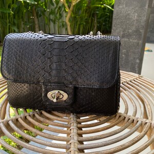 以下が含まれることがあります： 蛇柄の黒い革製のハンドバッグ。シルバーの留め具とチェーンストラップが付いている。
