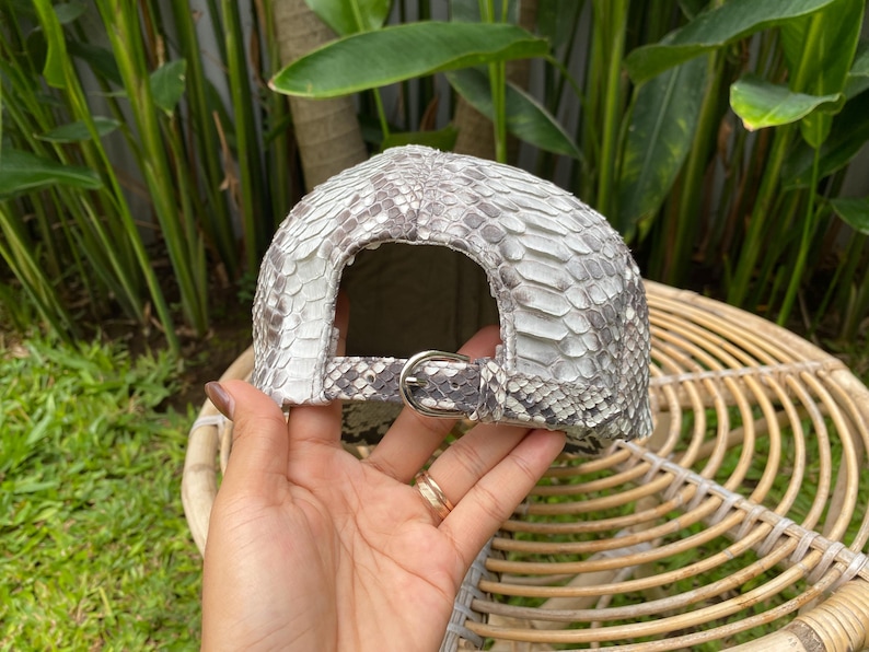 May include: A gray and white snakeskin baseball cap with a silver buckle closure.