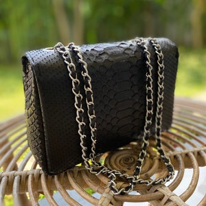 以下が含まれることがあります： 銀色のチェーンストラップが付いた黒いヘビ革のハンドバッグ。バッグは籐製の椅子の上に置かれています。