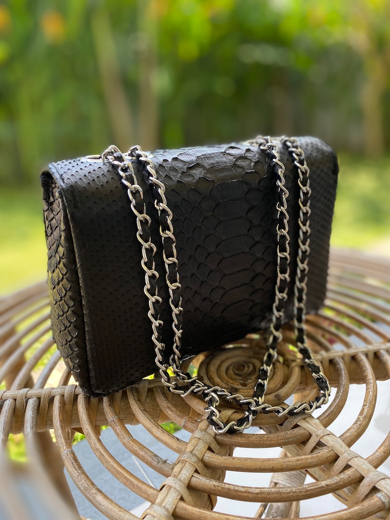 以下が含まれることがあります： 銀色のチェーンストラップが付いた黒いヘビ革のハンドバッグ。バッグは籐製の椅子の上に置かれています。