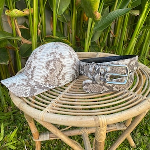 May include: A brown and white snake skin patterned baseball cap and belt set. The cap has a curved brim and the belt has a silver buckle.