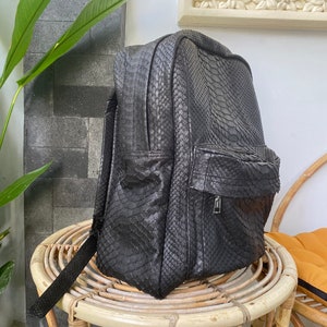 May include: A black leather backpack with a snakeskin pattern. The backpack is sitting on a wicker chair.