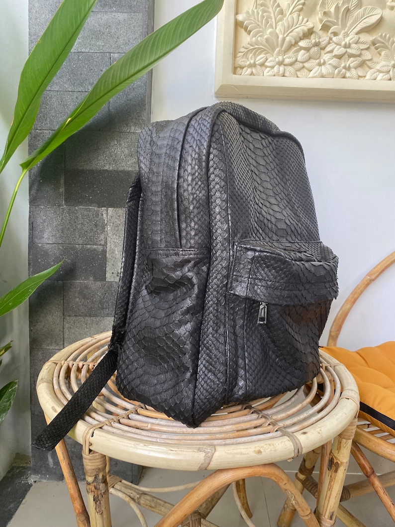 May include: A black leather backpack with a snakeskin pattern. The backpack is sitting on a wicker chair.