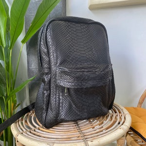 May include: A black leather backpack with a snakeskin pattern. The backpack is sitting on a wicker chair.