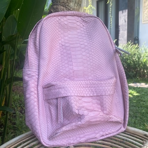 May include: A pink snakeskin backpack with a zippered front pocket. The backpack is made of genuine leather and has a unique, textured design.