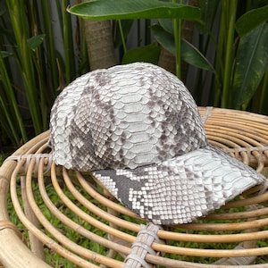 May include: A white and gray snakeskin baseball cap with a curved brim. The cap is sitting on a woven wicker surface.