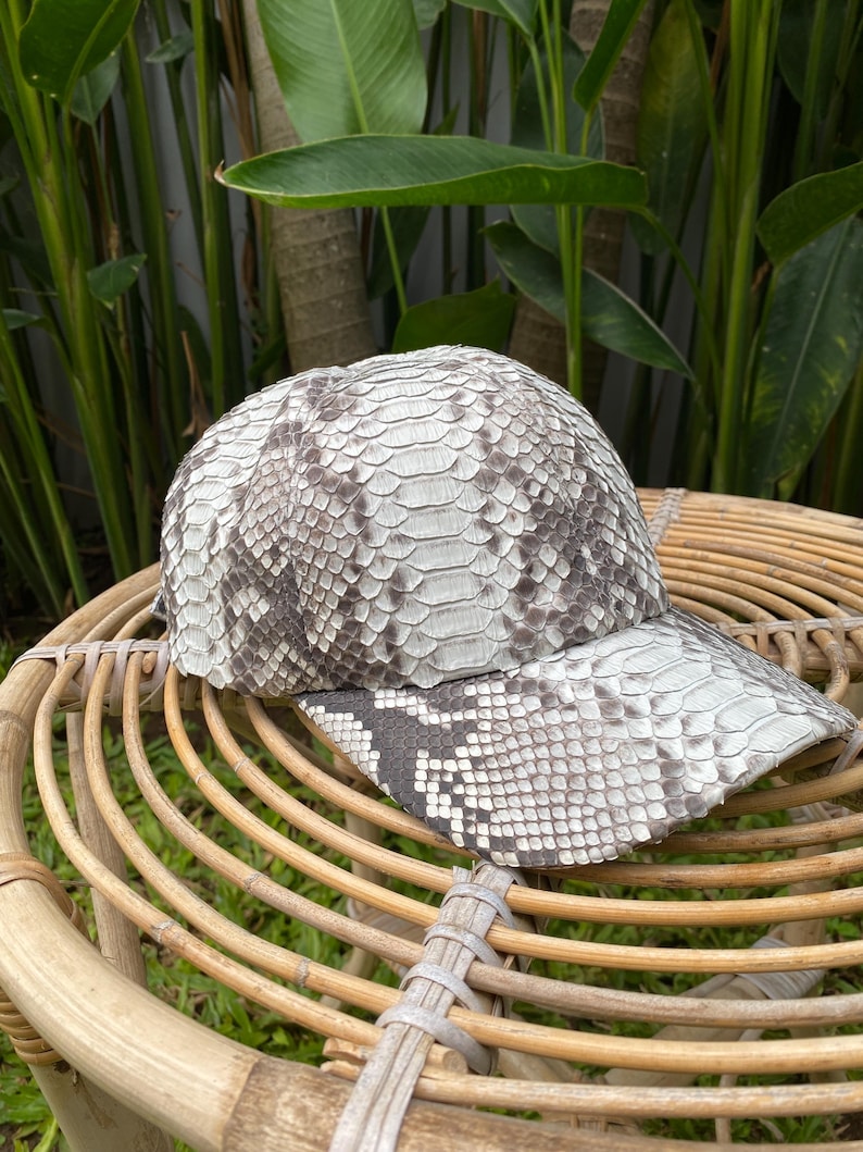May include: A white and gray snakeskin baseball cap with a curved brim. The cap is sitting on a woven wicker surface.