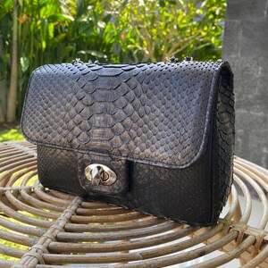 以下が含まれることがあります： シルバーのチェーンストラップとシルバーの留め金が付いた、黒いヘビ革のショルダーバッグです。