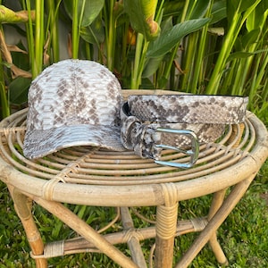 May include: A brown snakeskin baseball cap and a matching snakeskin belt with a silver buckle. The items are sitting on a woven wicker table.
