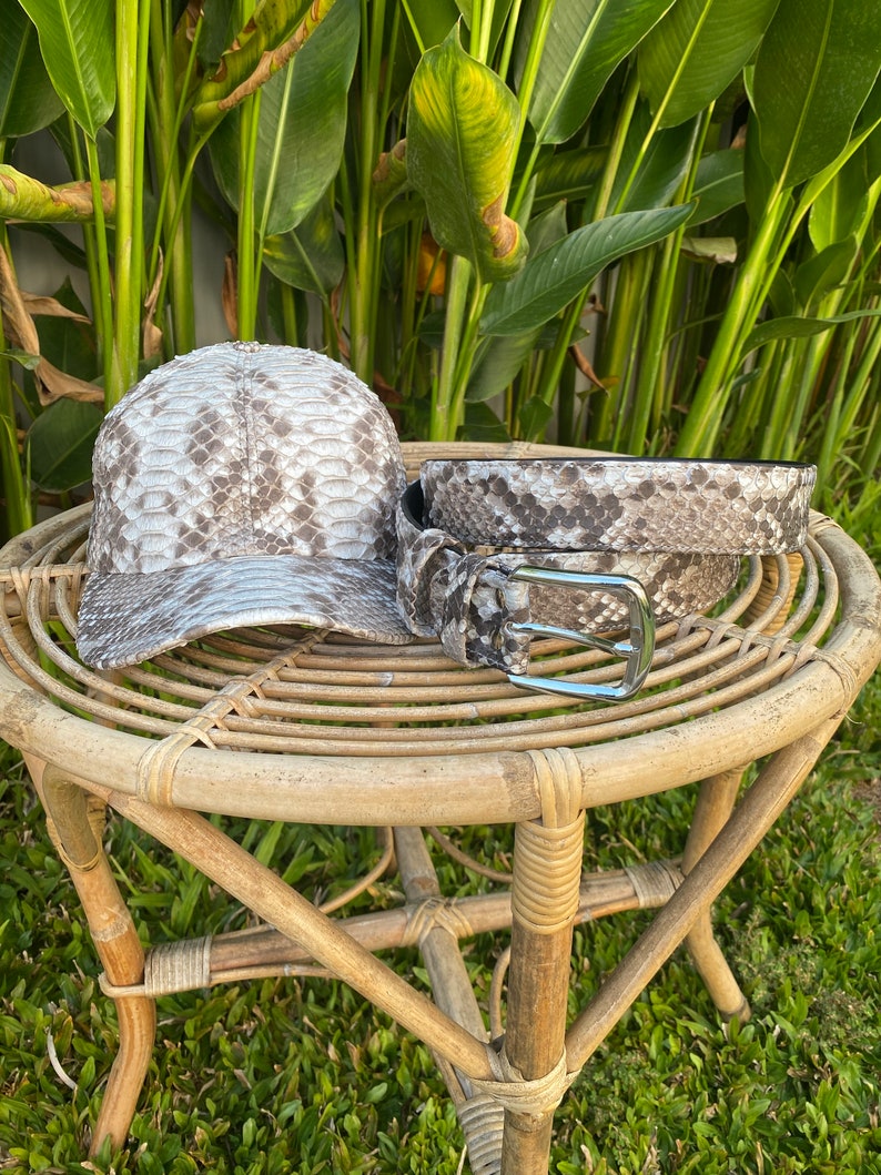 May include: A brown snakeskin baseball cap and a matching snakeskin belt with a silver buckle. The items are sitting on a woven wicker table.