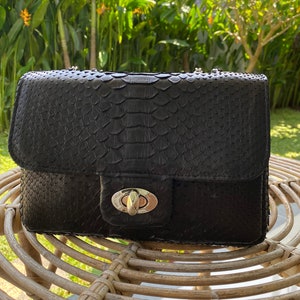 以下が含まれることがあります： シルバーのチェーンストラップとシルバーの留め具が付いた、黒いヘビ革のショルダーバッグです。