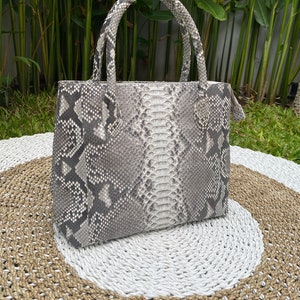May include: A white and grey snakeskin handbag with a top handle. The bag is sitting on a woven rug.