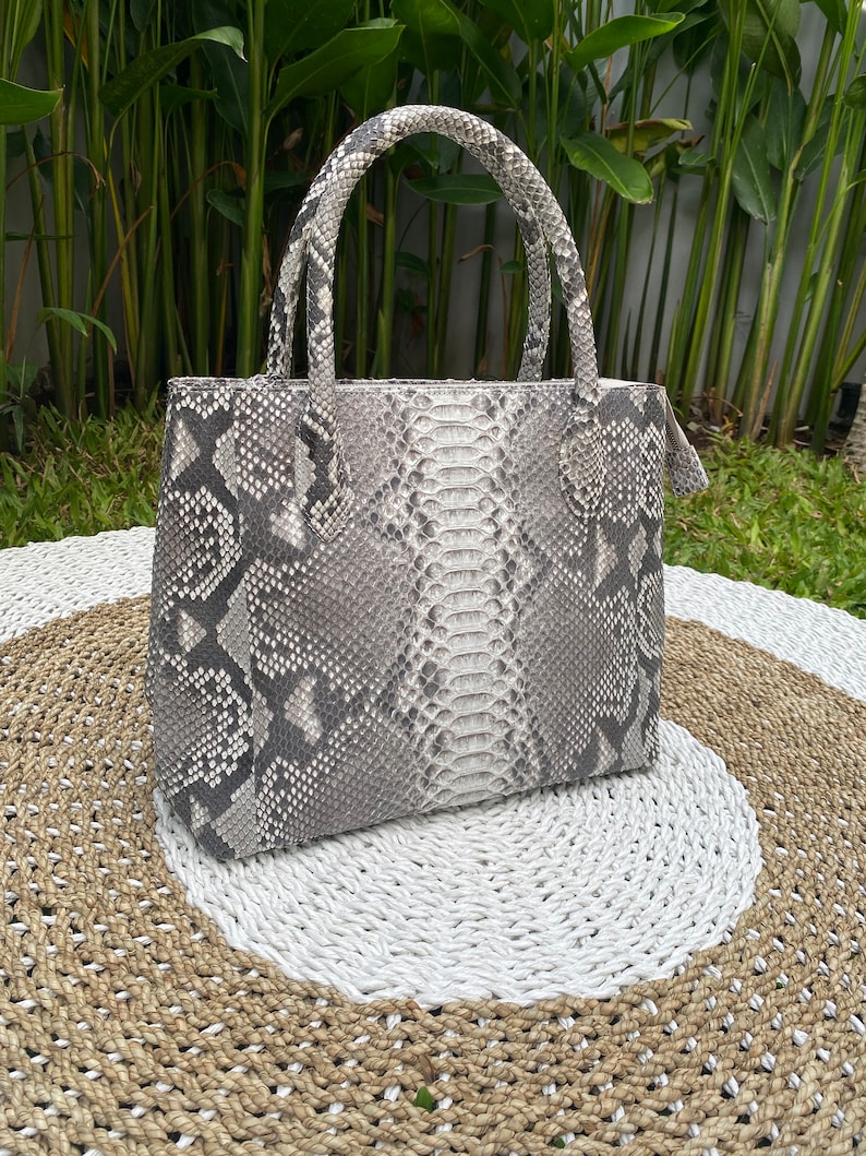 May include: A white and grey snakeskin handbag with a top handle. The bag is sitting on a woven rug.