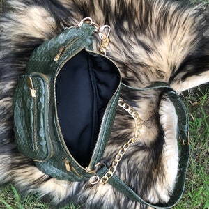 Puede incluir: Una ri&ntilde;onera de piel de serpiente verde con una correa de cadena dorada. La bolsa tiene varios compartimentos con cremallera y est&aacute; abierta para mostrar el forro negro.