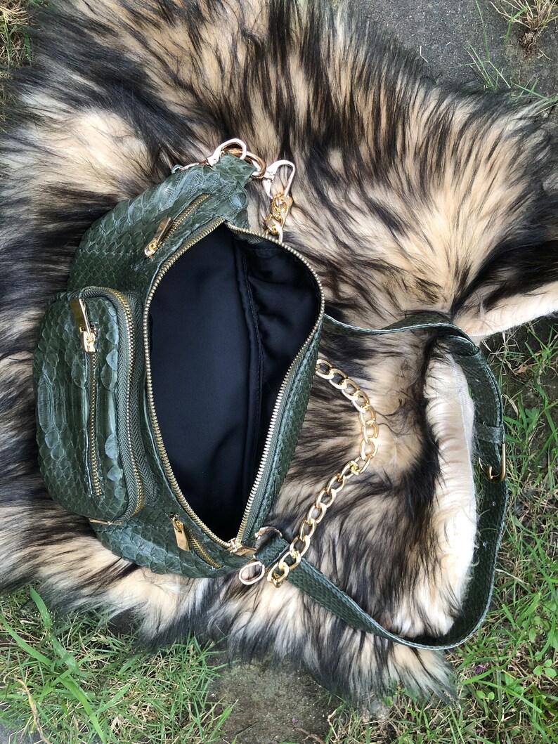 Puede incluir: Una ri&ntilde;onera de piel de serpiente verde con una correa de cadena dorada. La bolsa tiene varios compartimentos con cremallera y est&aacute; abierta para mostrar el forro negro.