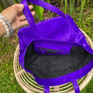 Puede incluir: Un bolso tote de cuero morado con estampado de piel de serpiente. El bolso tiene dos asas superiores y un forro de tela negro.