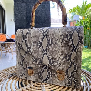 Pode incluir: Uma bolsa de mão de pele de cobra marrom e preta com alça de bambu. A bolsa tem dois fechos dourados na frente.