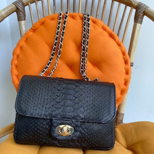 May include: A black snakeskin handbag with a gold chain strap and a gold clasp. The bag is sitting on a bright orange cushion.