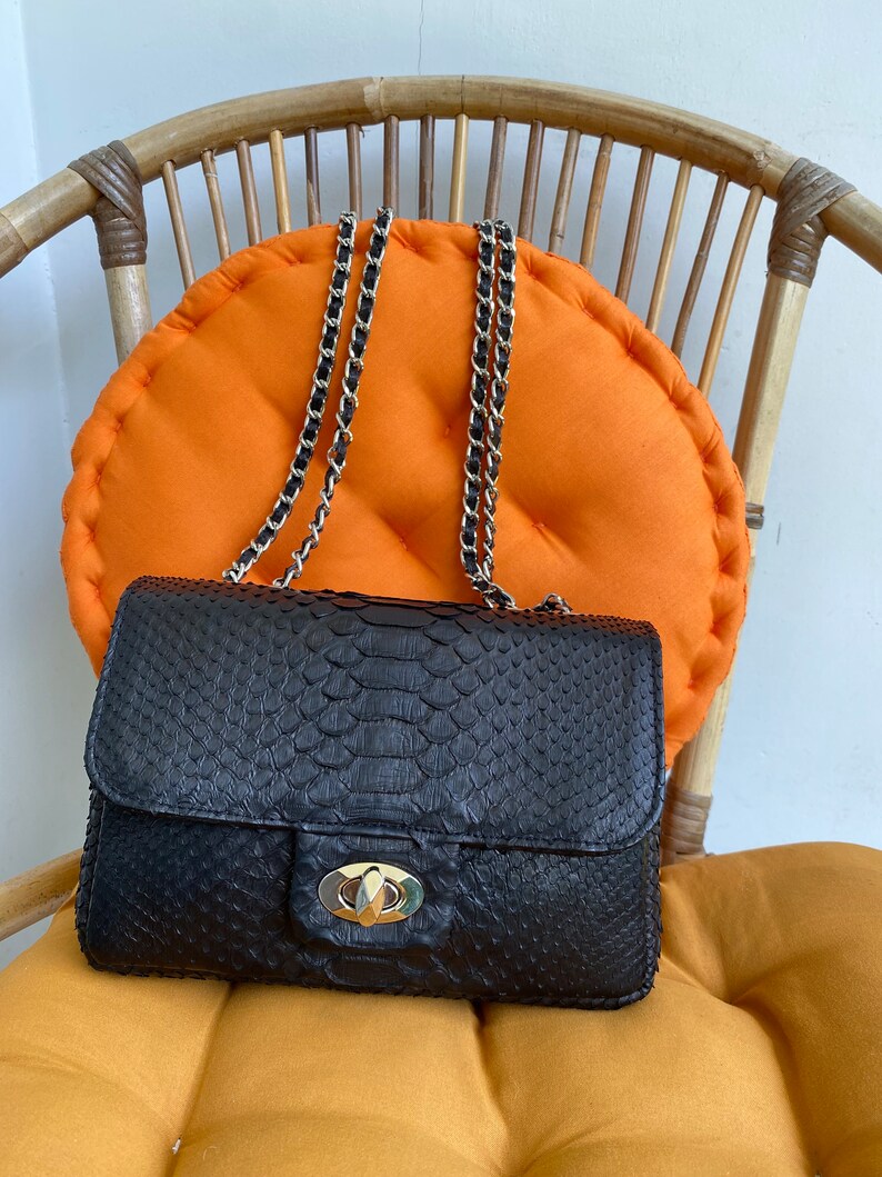 以下が含まれることがあります： 金色のチェーンストラップと金色の留め具が付いた黒いヘビ革のハンドバッグ。バッグは明るいオレンジ色のクッションの上に置かれています。