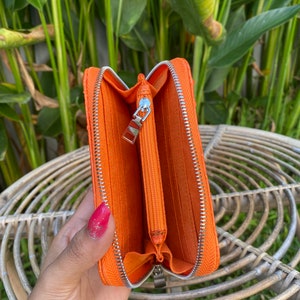 May include: Orange leather zippered wallet with a silver zipper. The wallet is open and the inside is lined with a soft orange fabric.