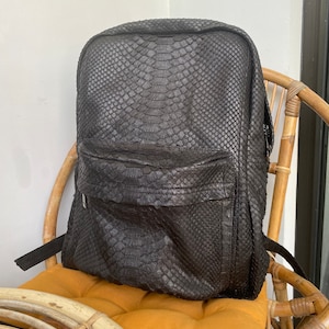 May include: A black leather backpack with a snakeskin pattern. The backpack is sitting on a wicker chair with a yellow cushion.