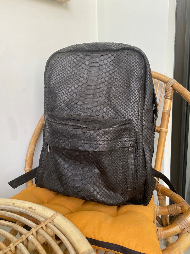 May include: A black leather backpack with a snakeskin pattern. The backpack is sitting on a wicker chair with a yellow cushion.