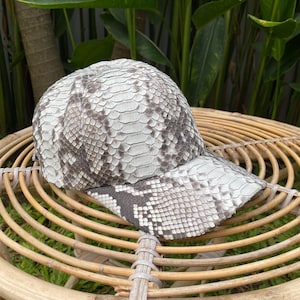 May include: A white and gray snakeskin baseball cap with a curved brim. The cap is sitting on a woven wicker surface.