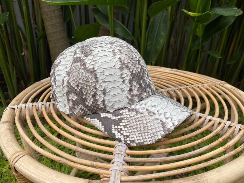 May include: A white and gray snakeskin baseball cap with a curved brim. The cap is sitting on a woven wicker surface.