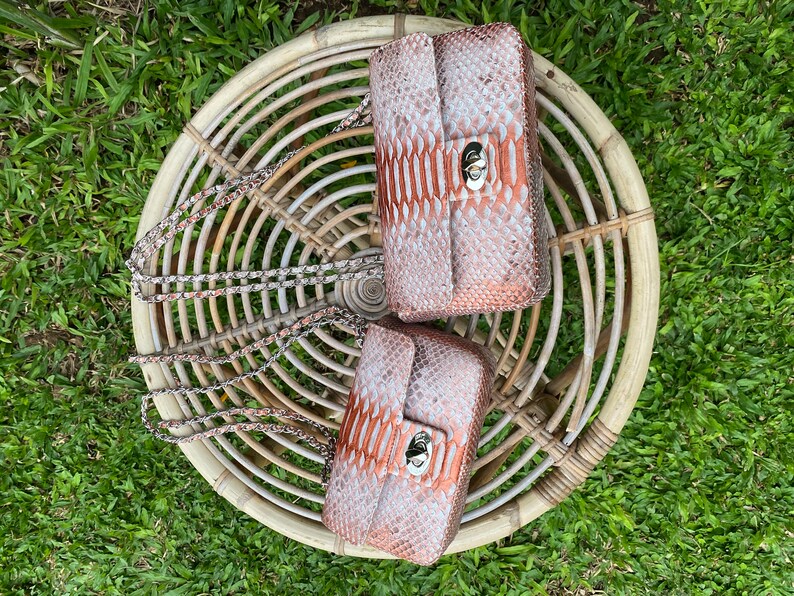 May include: Two small, brown and white snakeskin purses with silver chains and clasps. The purses are sitting on a woven wicker chair.