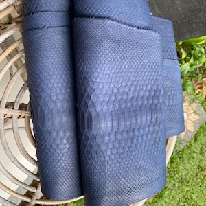 Pode incluir: Close-up de vários rolos de couro de pele de cobra azul marinho. O couro tem um padrão escamoso texturizado e é enrolado firmemente. Os rolos são dispostos em uma cadeira de vime, com grama verde visível ao fundo. O couro parece ser de alta qualidade.