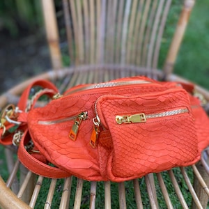 Op de afbeelding: Een oranje heuptas met een textuur die op slangenleer lijkt. De tas heeft meerdere compartimenten met ritssluiting en goudkleurige hardware. De tas ligt op een houten stoel.