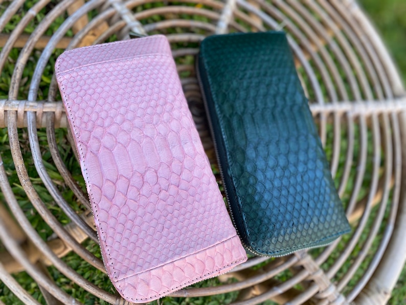 Peut inclure: Deux portefeuilles &agrave; fermeture &eacute;clair en peau de serpent. L'un est rose et l'autre vert. Les portefeuilles sont pos&eacute;s sur une surface en osier tress&eacute;.