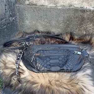 Könnte beinhalten: Eine schwarze Leder-Bauchtasche mit einem silbernen Kettenriemen und einem Schlangenhautmuster. Die Tasche hat mehrere Fächer und einen Reißverschluss.