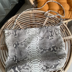 Könnte beinhalten: Eine weiße und graue Schlangenleder-Handtasche mit einem langen Riemen. Die Tasche liegt auf einem Korbstuhl.