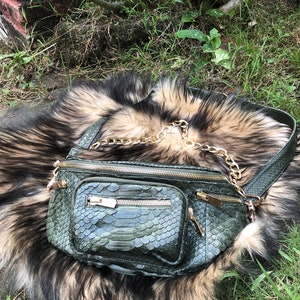 Puede incluir: Una ri&ntilde;onera de piel de serpiente verde con una correa de cadena dorada. La bolsa tiene varios compartimentos con cremallera y est&aacute; sobre una alfombra de piel suave, marr&oacute;n y blanca.