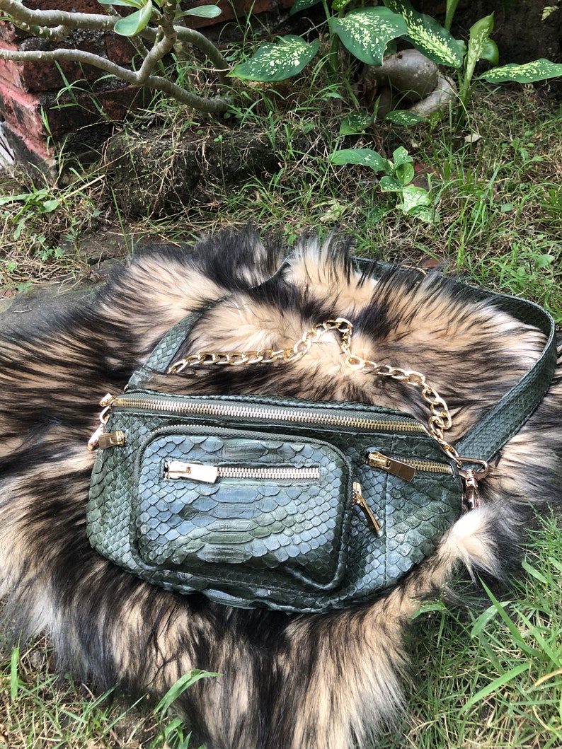 Puede incluir: Una ri&ntilde;onera de piel de serpiente verde con una correa de cadena dorada. La bolsa tiene varios compartimentos con cremallera y est&aacute; sobre una alfombra de piel suave, marr&oacute;n y blanca.