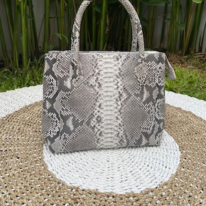 May include: A grey and white snakeskin handbag with a top handle. The bag is sitting on a woven rug.