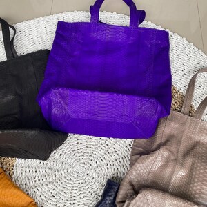 Puede incluir: Cinco bolsos tote de cuero en varios colores, incluyendo morado, negro, marr&oacute;n, naranja y azul. Los bolsos est&aacute;n hechos de cuero texturizado con un patr&oacute;n de piel de serpiente. Los bolsos est&aacute;n dispuestos sobre una superficie blanca tejida.