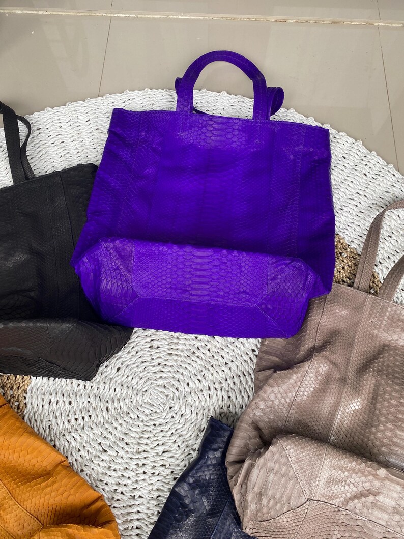 Puede incluir: Cinco bolsos tote de cuero en varios colores, incluyendo morado, negro, marr&oacute;n, naranja y azul. Los bolsos est&aacute;n hechos de cuero texturizado con un patr&oacute;n de piel de serpiente. Los bolsos est&aacute;n dispuestos sobre una superficie blanca tejida.