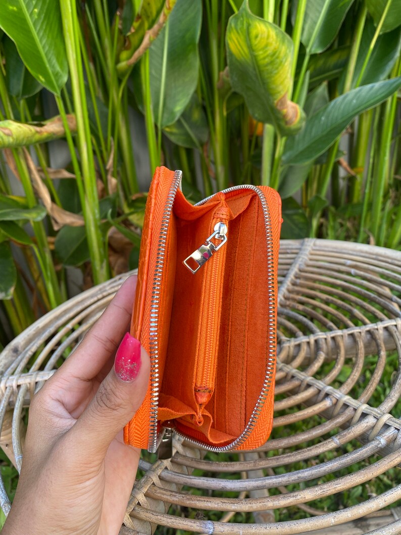 May include: Orange leather zippered wallet with a silver zipper. The wallet is open and the inside is lined with orange fabric.