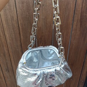 May include: Silver clutch bag with a chain strap. The bag has a textured, metallic finish and a silver chain strap. The interior of the bag is also silver. The bag is open, revealing the inside.