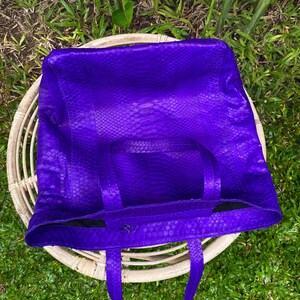 Puede incluir: Un bolso tote de cuero morado con estampado de piel de serpiente. El bolso tiene dos asas superiores y una sola correa.