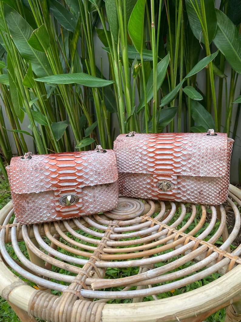 May include: Two small, light pink snakeskin purses with silver hardware. The purses are sitting on a woven wicker stool.