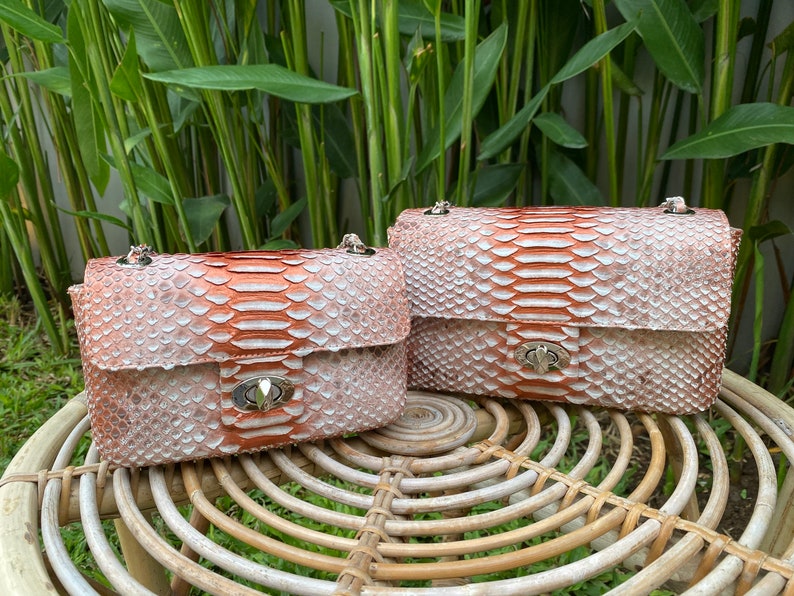 May include: Two small, light pink and orange snakeskin purses with silver hardware and chain straps. The purses are sitting on a woven wicker table.
