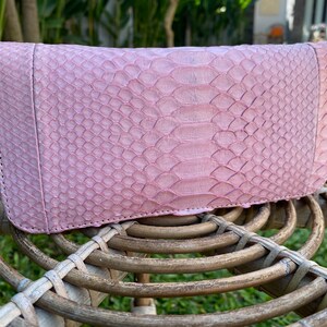 Peut inclure: Un portefeuille en cuir de serpent rose avec une fermeture &agrave; glissi&egrave;re. Le portefeuille est pos&eacute; sur une surface en osier tress&eacute;.