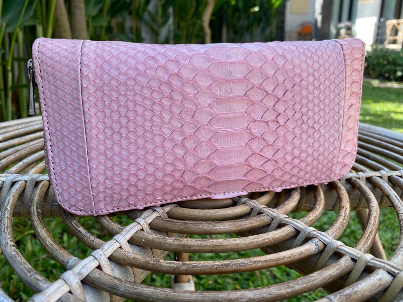 Peut inclure: Un portefeuille en cuir de serpent rose avec une fermeture &agrave; glissi&egrave;re. Le portefeuille est pos&eacute; sur une surface en osier tress&eacute;.