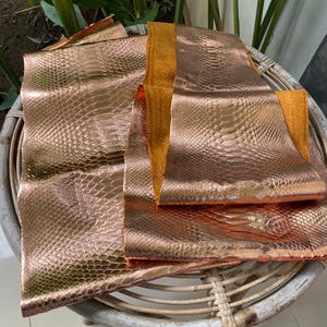 May include: Two pieces of rose gold snakeskin leather with a textured pattern. The leather is folded and stacked on a woven wicker chair.